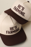 Two cream and brown "He's Farming." embroidered trucker snapback hats stacked together, featuring a curved brown brim and bold brown embroidered lettering.