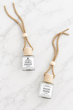 Two small glass diffuser bottles with white labels and light wood caps, hanging from braided gold-and-white cords with wooden beads, arranged on a white marble surface. One label reads NOBLE KNIGHT and the other reads BOUJE HEIFER.