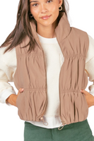 Woman wearing a mocha colored mini puffer style vest over a white long sleeve shirt. Vest has a front zipper closure and a drawstring adjustable waist.