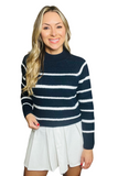 woman wearing a navy high-neck sweater with white stripes. Sweater is paired with a white pleated skirt 