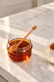 A glass jar filled with amber honey sits on a white marble countertop with a wooden honey dipper resting inside. A white teacup with gold trim is partially visible on the right, and warm sunlight casts shadows across the counter.