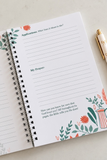 An open spiral-bound journal or planner page with lined writing areas and floral illustrations in teal, coral, and green, photographed on a light surface with a gold pen nearby.