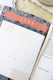 An open planner showing the month of June with a floral-patterned cover, photographed on a light tabletop.