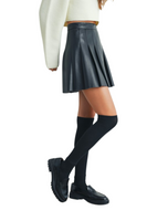Woman wearing a black pleated faux leather mini skirt styled with a cropped cream sweater, knee-high black socks, and black loafers for a chic street style look.