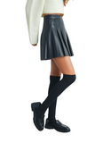 Woman wearing a black pleated faux leather mini skirt styled with a cropped cream sweater, knee-high black socks, and black loafers for a chic street style look.