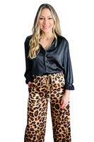 woman wearing a black sating button down collared shirt with the sleeves rolled up, the shirt is tucked into a pair os satin like drawstring leopard pants. Her hand is in the pocket 