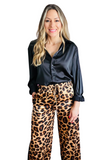 woman wearing a black sating button down collared shirt with the sleeves rolled up, the shirt is tucked into a pair os satin like drawstring leopard pants. Her hand is in the pocket 