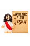 A small figurine of Jesus with brown hair and beard, a white robe, and a red sash stands beside a rectangular light wood sign engraved with the phrase 'EVERYONE NEEDS A LITTLE Jesus' in black lettering, on a plain white background.