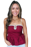 A smiling woman wearing a maroon camisole top with white trim and white spaghetti straps, paired with light blue jeans and a gold chain necklace, against a plain white background.