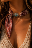 A close-up of a woman wearing a western-style bandana choker necklace. The necklace features a maroon paisley patterned fabric tied in a knot, with a central silver-toned concho-style clasp adorned with a turquoise stone.