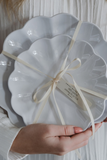 Two scalloped-edge ceramic plates in a glossy white finish, wrapped together with a cream ribbon and held by a person's hands against a soft white textured background.