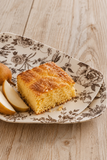 A square slice of golden-brown baked cake or bread on a beige floral ceramic plate, with pear slices beside it, set on a light wood tabletop.