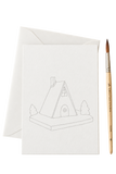 A white textured card with a simple line drawing of a house, shown with a matching white envelope behind it and a fine-tip paintbrush to the right on a clean white background.