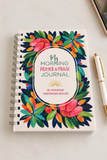 A spiral-bound morning prayer and praise journal with a colorful floral wreath design on the cover, placed on a light surface next to a pen.