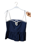 A navy blue camisole top with white crochet trim along the neckline and thin shoulder straps, hanging on a white hanger against a white background. A hand is holding the hanger on the right side.