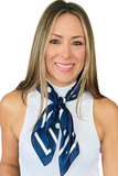A smiling woman with blonde-highlighted hair wearing a white ribbed sleeveless top and a navy blue silk scarf with white polka dots and abstract white lines, set against a plain white background.