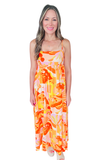 Woman wearing The Sunset Spritz Maxi Dress featuring a colorful tropical beach print with orange, pink, and yellow tones, square neckline, adjustable spaghetti straps, empire waist, and flowy lined maxi silhouette.