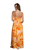 Back view of woman wearing The Sunset Spritz Maxi Dress featuring a colorful tropical beach print in orange, pink, and yellow tones with adjustable spaghetti straps, smocked back, empire waist, and flowy lined maxi skirt.