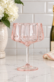 A pink fluted stemware glass with a wide bowl and a slender stem, set on a white marble countertop with a white subway tile backsplash, white flowers in a vase, and a bottle of wine in the background.