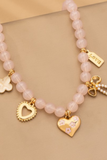 Close-up of pink beaded charm necklace with gold heart, bow, clover, and floral details, highlighting feminine charm accents on a neutral background