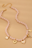 Pink beaded charm necklace with gold heart, bow, clover, and floral charms, featuring a delicate feminine design on a soft neutral background