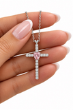 A silver cross pendant necklace with a central pink heart-shaped gemstone and surrounding clear stones, held in a hand with manicured nails against a white background.