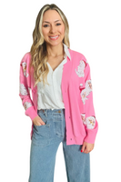 Woman wearing a pink button down cardigan with sequin Santa heads on the front over a white button down shirt. Cardigan is paired with light denim pants