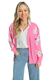 Woman wearing a pink button down cardigan with sequin Santa heads on the front over a white button down shirt. Cardigan is paired with light denim pants