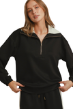 A woman modeling a black long-sleeve quarter-zip sweatshirt with a contrasting cream collar, paired with black track pants featuring a white side stripe, against a plain white background.