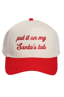 Cream and red Christmas trucker hat with a snapback closure, featuring embroidered red script text on the front that reads, “put it on my Santa’s tab.”