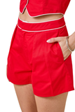 A close-up of a model wearing bright red shorts with white piping trim, showing the side pocket with a hand inside and a silver bangle bracelet.