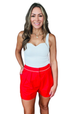 A woman modeling a light blue ribbed tank top and bright red shorts with a thin white waistband, set against a plain white background.