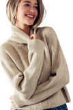 Woman wearing an oatmeal colored ribbed turtleneck sweater playfully pulling on the neckline