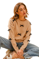 woman wearing a beige crew neck sweater with black ribbon appliqué. It is paired with cuffed light denim pants and sandals. 