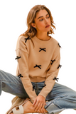 woman wearing a beige crew neck sweater with black ribbon appliqué. It is paired with cuffed light denim pants and sandals. 