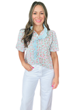 Woman wearing a lightweight blue floral blouse with pink floral print, puff sleeves, functional button front, and eyelet ruffle neckline detail tucked in to a pair of white wide leg pants.