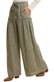 A person wearing loose, wide-leg drawstring pants with a small-scale floral or mottled print in muted green, beige, and gray tones, paired with a white top and brown shoes against a plain white background.