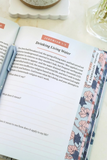 An open Bible or prayer journal showing Jeremiah 1:2 with the heading 'Drinking Living Water,' alongside a floral bookmark with pink, blue, and gray flowers. The pages are photographed on a light surface with a small dish and glass in the background.
