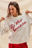 Model wearing a white Tis The Season Sequin Sweater with red sequin lettering, styled with statement earrings for a festive holiday outfit.