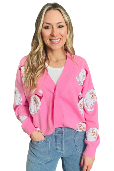 Woman wearing a pink button down cardigan with sequin Santa heads on the front. Cardigan is paired with light denim pants