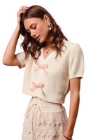 woman wearing a ivory knit textured v-neck short sleeve sweater with 2 light pink velvety bows on the front center. Sweater is paired with ivory lace drawstring bottoms 