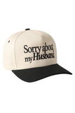 Side view of a cream and black trucker hat with black embroidered text reading “Sorry about my husband” on the front panel.