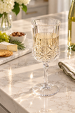 A clear cut-crystal wine glass filled with white wine, set on a light marble countertop in a bright kitchen setting with cheese, crackers, grapes, and a wine bottle nearby.