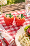 Two strawberry-shaped salt and pepper shakers with smiling faces and small white flowers on top, placed on a red-and-white checkered tablecloth beside a plate of food in an outdoor dining setting.