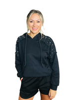 woman wearing a black hoodie with studded details on the shoulders. There is also a raw hemline on the shoulders.  Hoodie has a kangaroo front pocket.