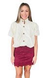 A woman modeling a cream puff-sleeve blouse with gold buttons and a burgundy mini skirt with a lace hem, set against a plain white background.