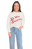 Model wearing a cream knit sweater with red sequin “Tis the Season” lettering, crew neckline, long sleeves, and a relaxed fit, styled with light wash jeans.