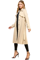 woman wearing a beige pleated trench coat. Coat is called has brown buttons down the front 