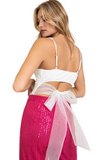 woman turned to her side looking back, wearing a white bodysuit with a cut out in the back and a tulle bow in the back. She is wearing hot pink sequin pants 