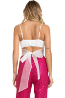 Back of a woman wearing a white bodysuit with a cut out and tie tulle bow detail. Bodysuit is a spaghetti strap with adjustable straps. She is wearing hot pink sequin pants 